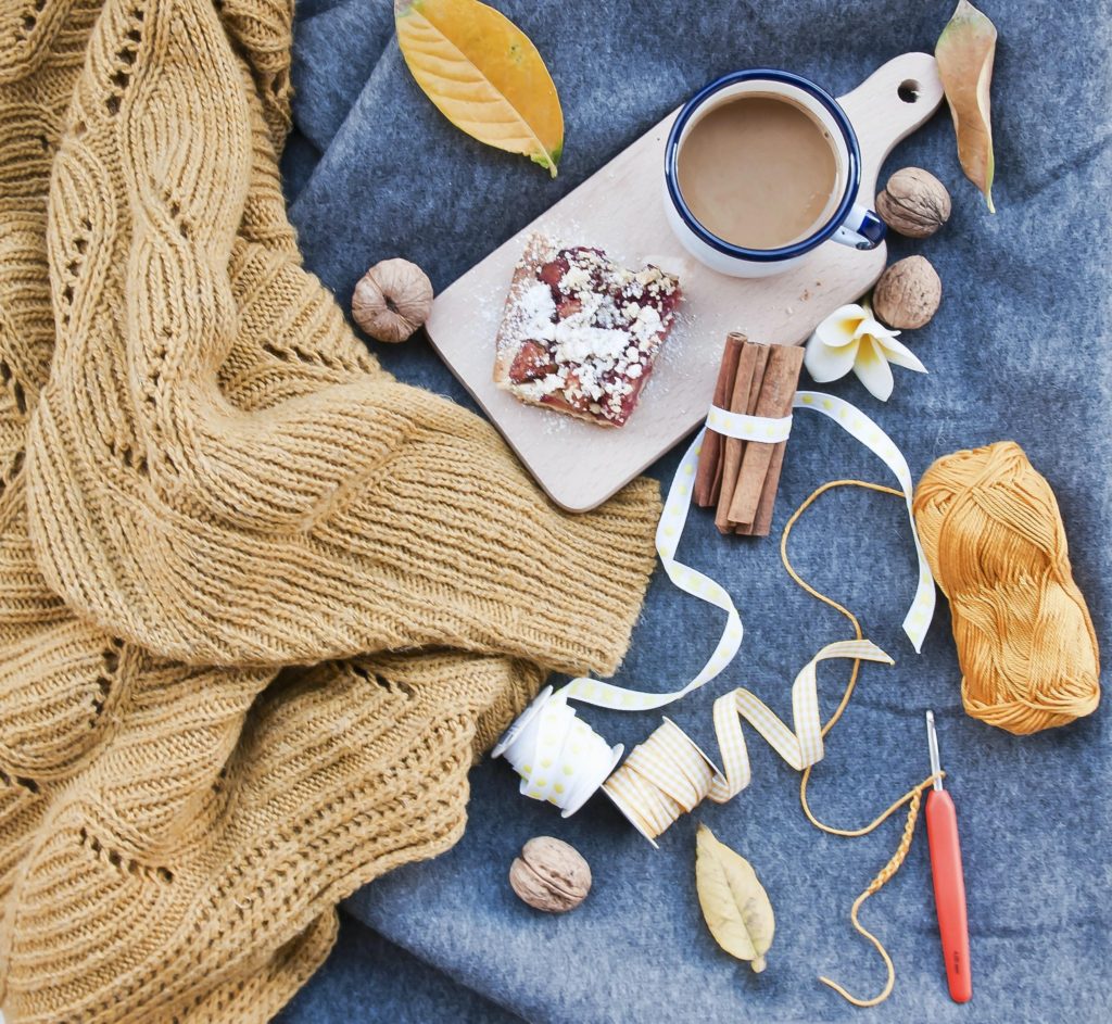 A collage of autumn craft supplies, along with a warm knitted jumper, ribbons, wool, cinnamon sticks, a cup of hot chocolate and a snack. 