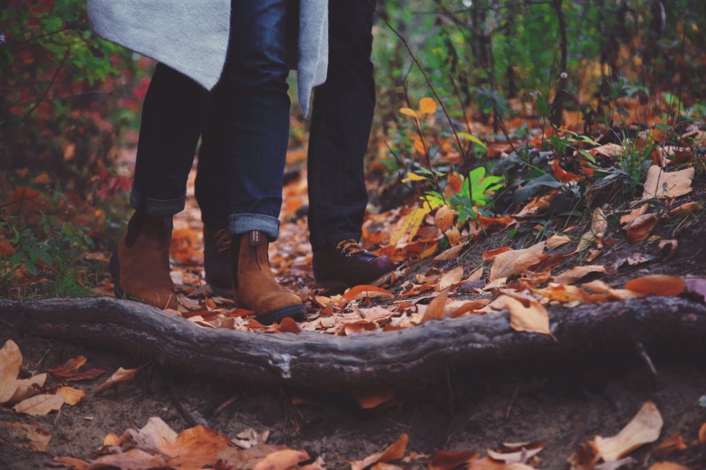 Two people stand in a forest, the photo showing their feet in hiking boots, surrounded by autumn leaves. 