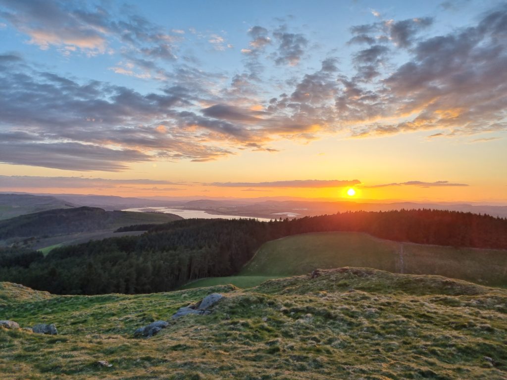 Sunset walk: A view from a hill summit with the sun setting on the right hand side beyond mountains and a river. 