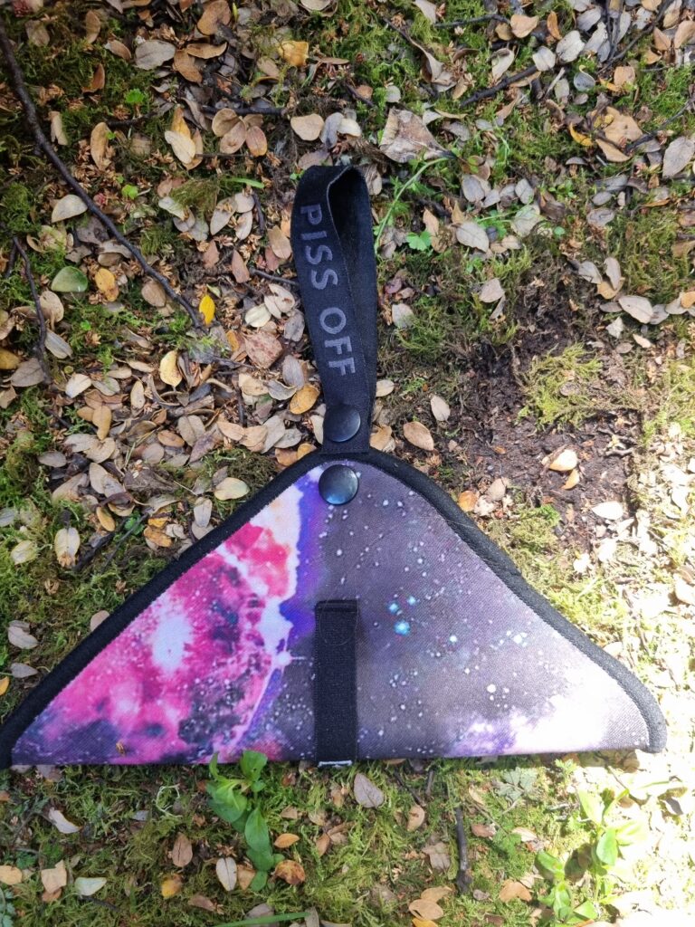 A picture of my pee cloth. It's pink and purple, with a stud hanger to hang it from my backpack, stamped with the words "piss off". The background is a leafy forest floor. 