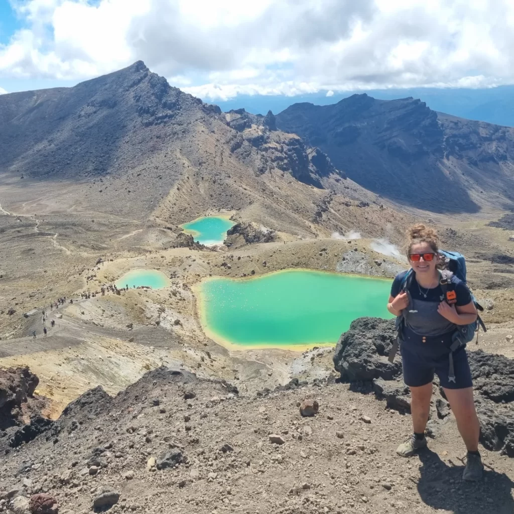 Image shows me standing and smiling, with the turquoise lakes of the Tongariro Crossing in the background, and whisps of smoke from the active volcanos decorating the ground below. 