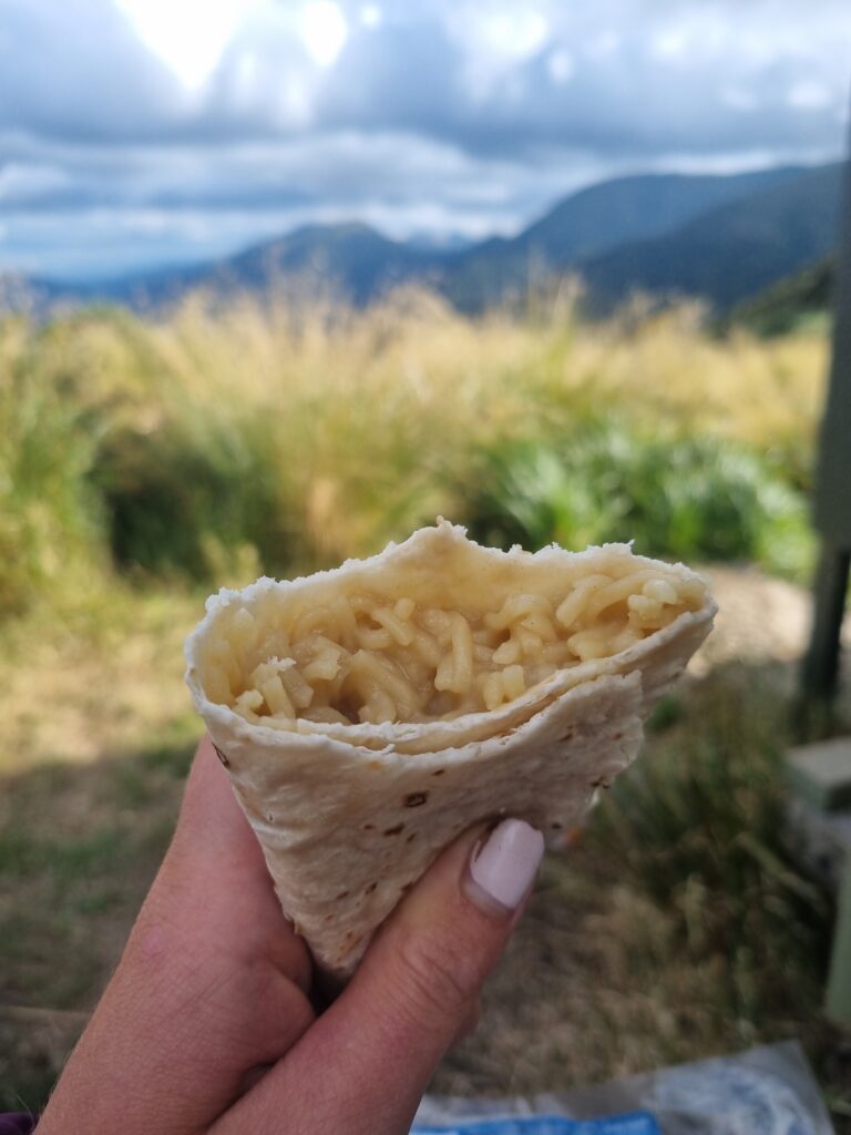 The image focuses on a white wrap in my hand, filled with noodles (and unseen mashed potato flakes). The blurry background is full of yellow tussock, green grasses and expansive mountain ranges. 