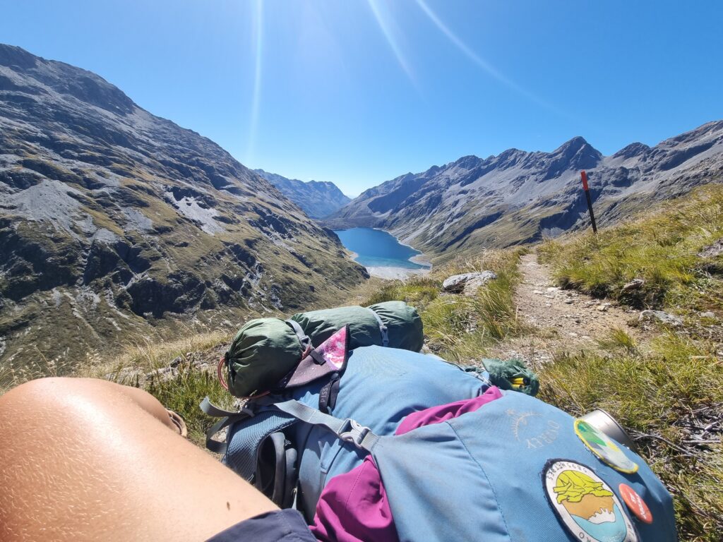 On a glorious sunny day, the aqua blue of Lake Constance mimicks the sky when viewed from the pass. My blue backpack covered in badges and gear sits next to the tanned (and tired) knee. One of the best views of Te Araroa. 